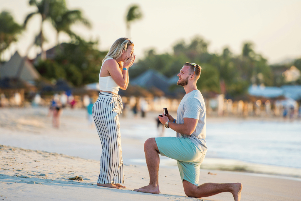 A man down on on knee proposing on a beach inThailand. Travel engagement ring. Download Tengah App