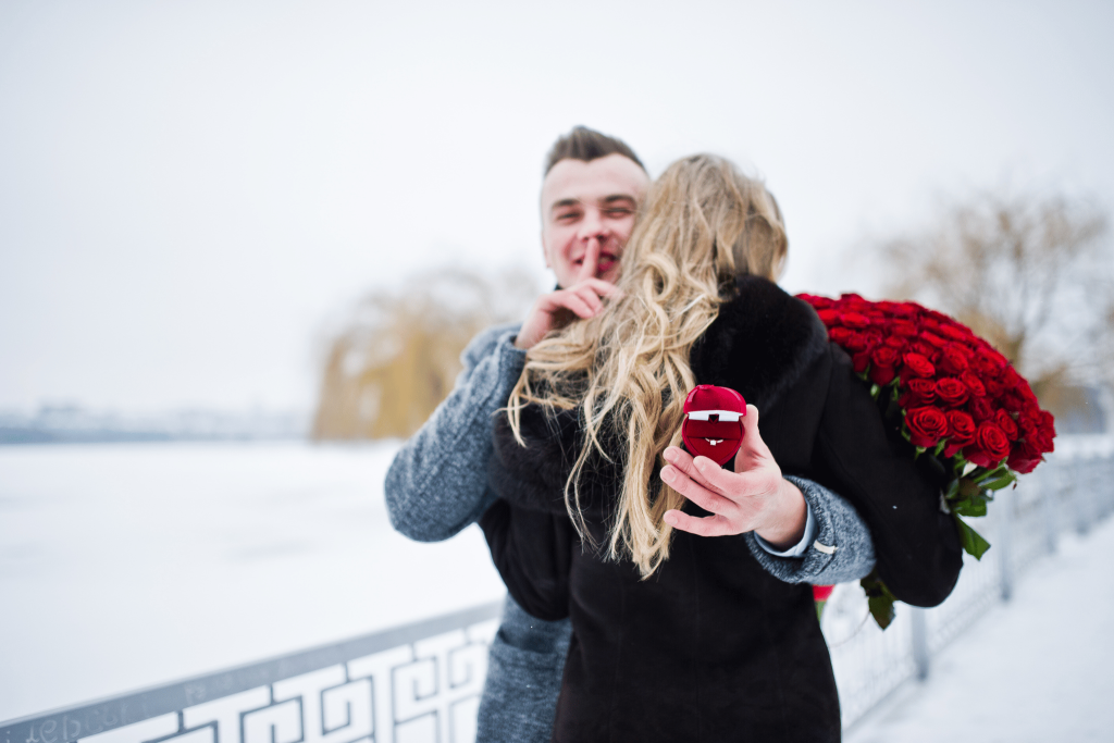 Man hugging his girlfriend with an engagement ring as a secret being held behind her back. Red roses in the snow. 