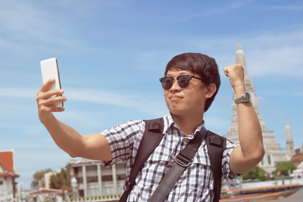 Asian guy posing in front of his phone camera taking a picture of him on holiday. Instagram versus reality. The stress of travel. Download Tengah